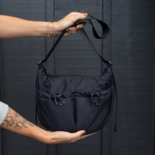 A person holding a black shoulder bag with two exterior cinch pockets and a longer crossbody strap.
